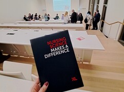 Hand hält Broschüre mit Titel Nursing Science makes a difference hoch im Hörsaal...